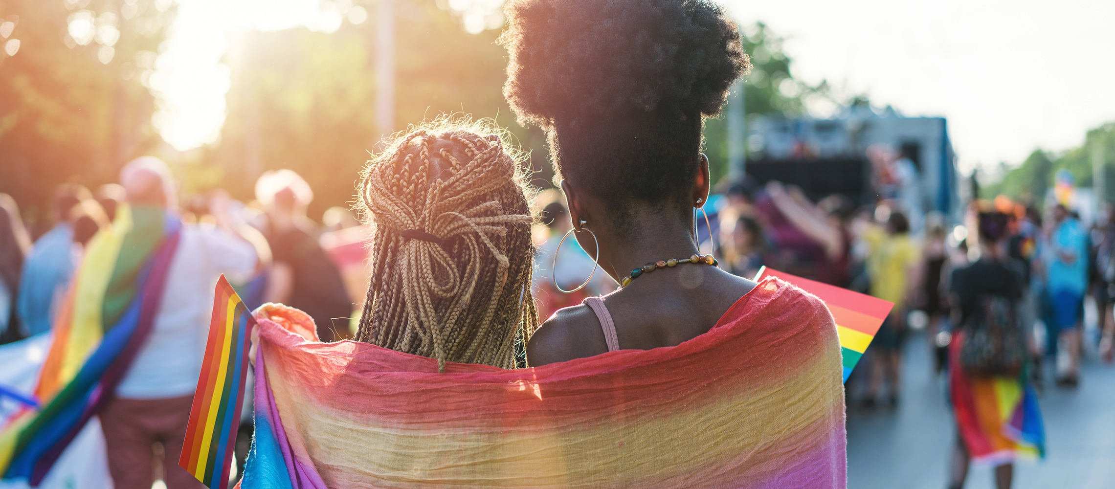 Two women standing at a Pride parade with a Pride Flag wrapped around their shoulders.
